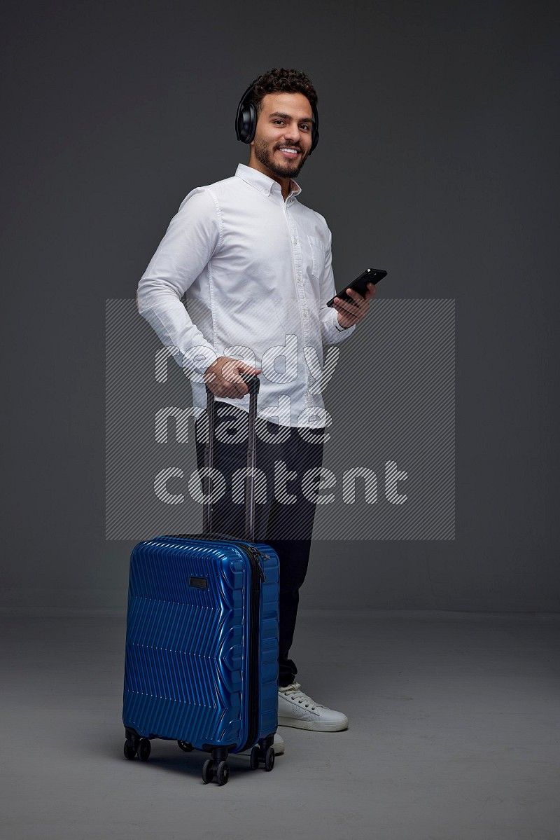 A man wearing smart casual and headphone holding luggage eye level on a gray background