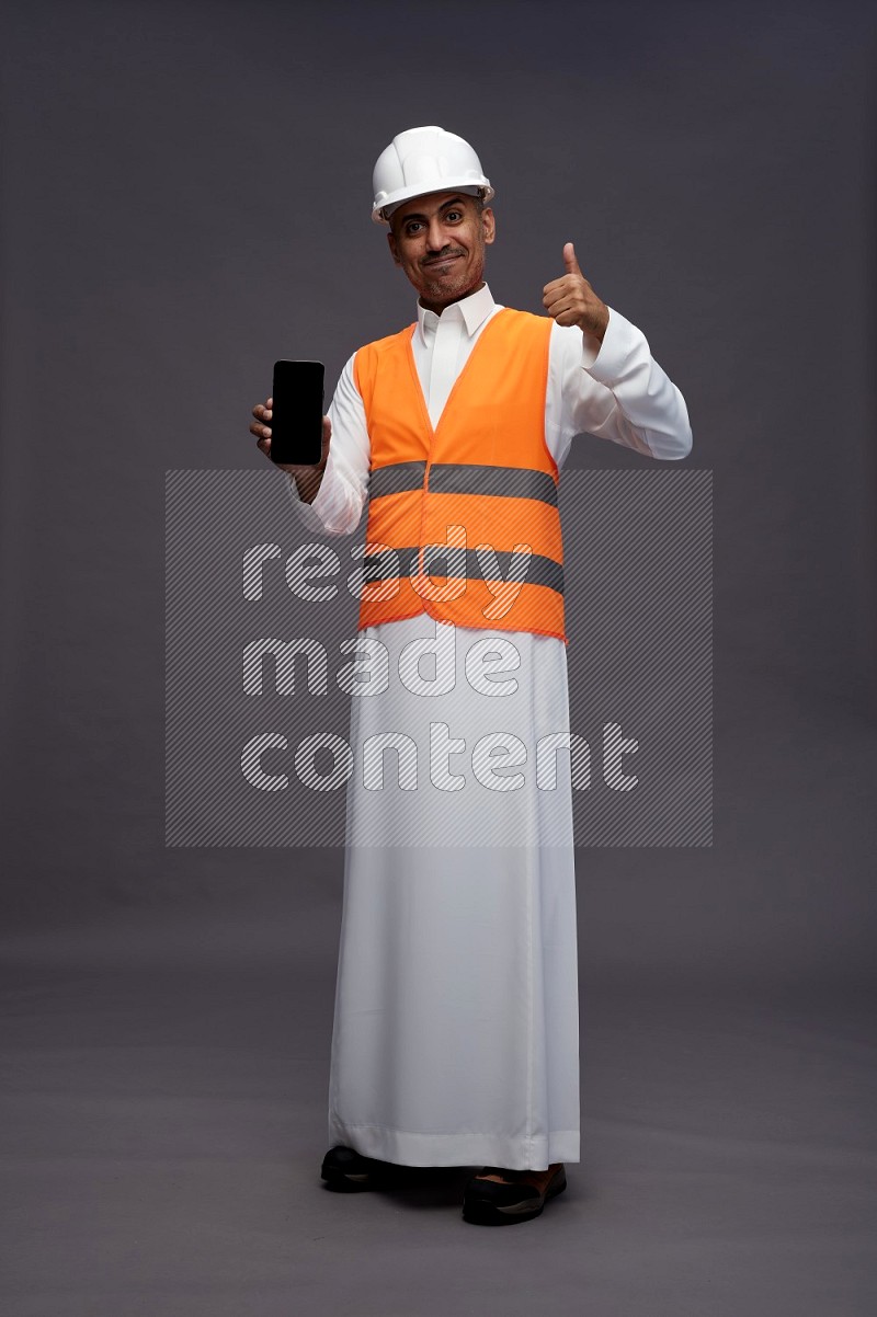 Saudi man wearing thob with engineer vest standing showing phone to camera on gray background