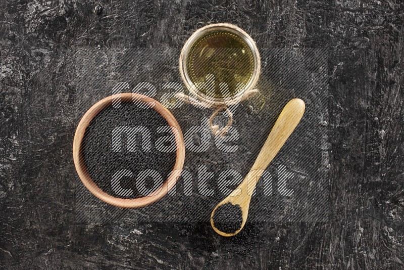 A wooden bowl and spoon full of black seeds and a glass jar of black seeds oil on a textured black flooring in different angles