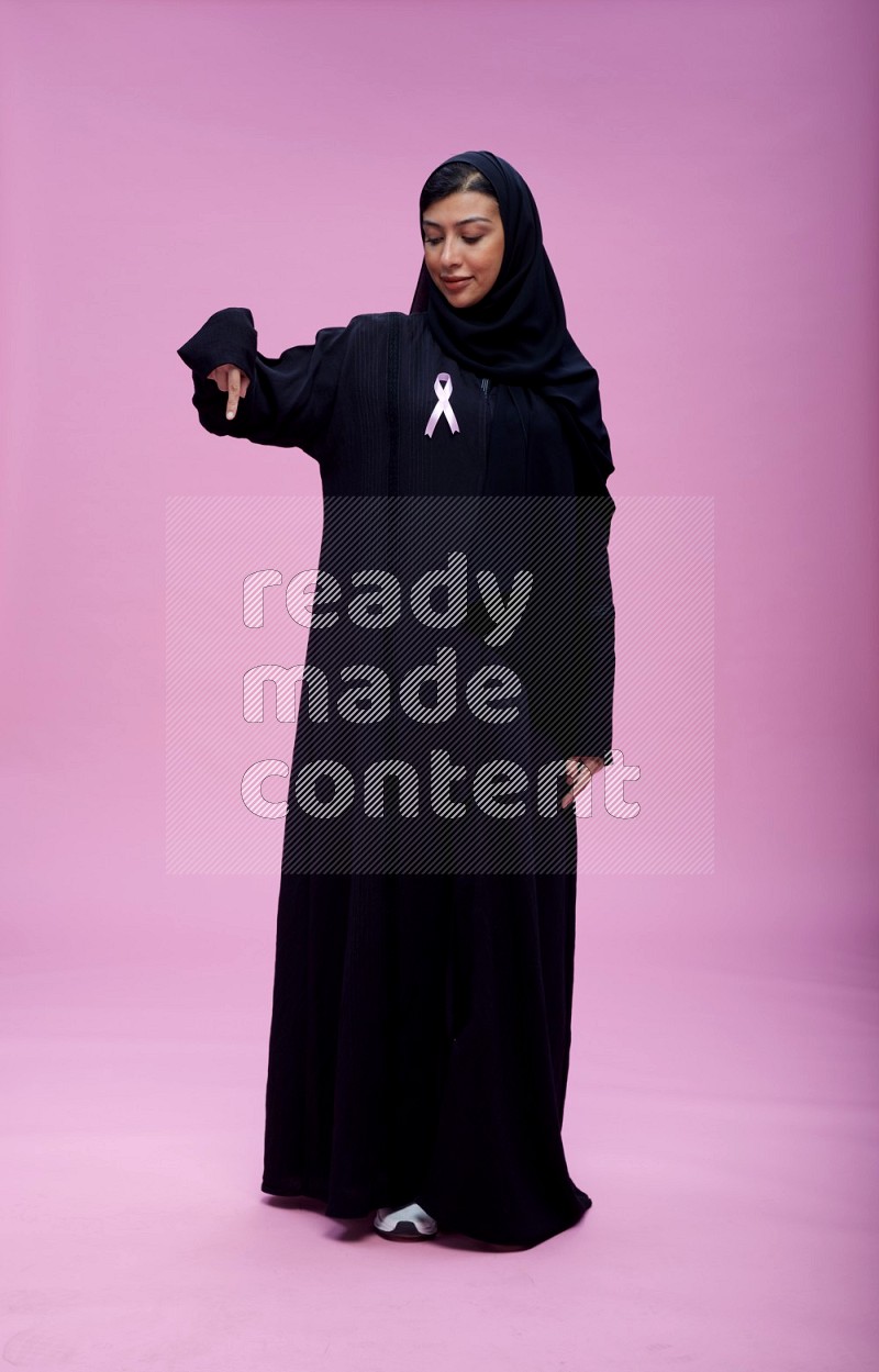 Saudi woman wearing pink ribbon on Abaya standing interacting with the camera on pink background