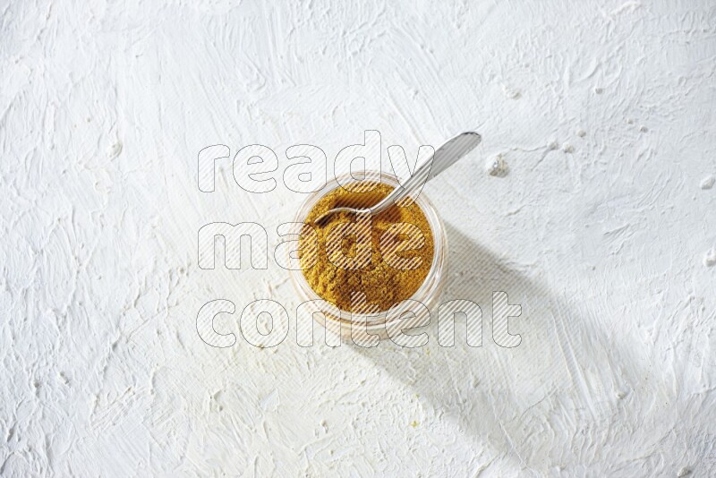 A glass jar and a metal spoon full of turmeric powder on a textured white flooring