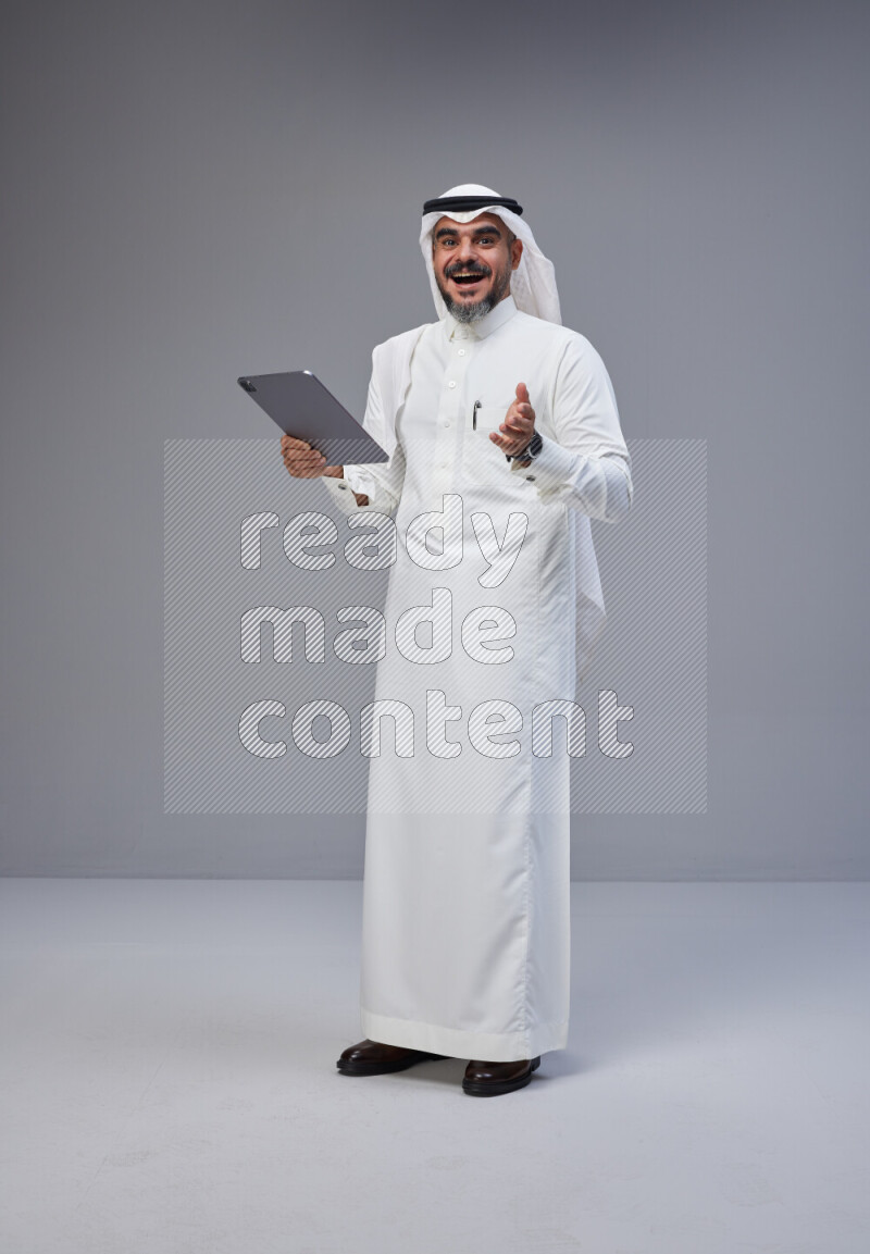Saudi man Wearing Thob and white Shomag standing working on tablet on Gray background