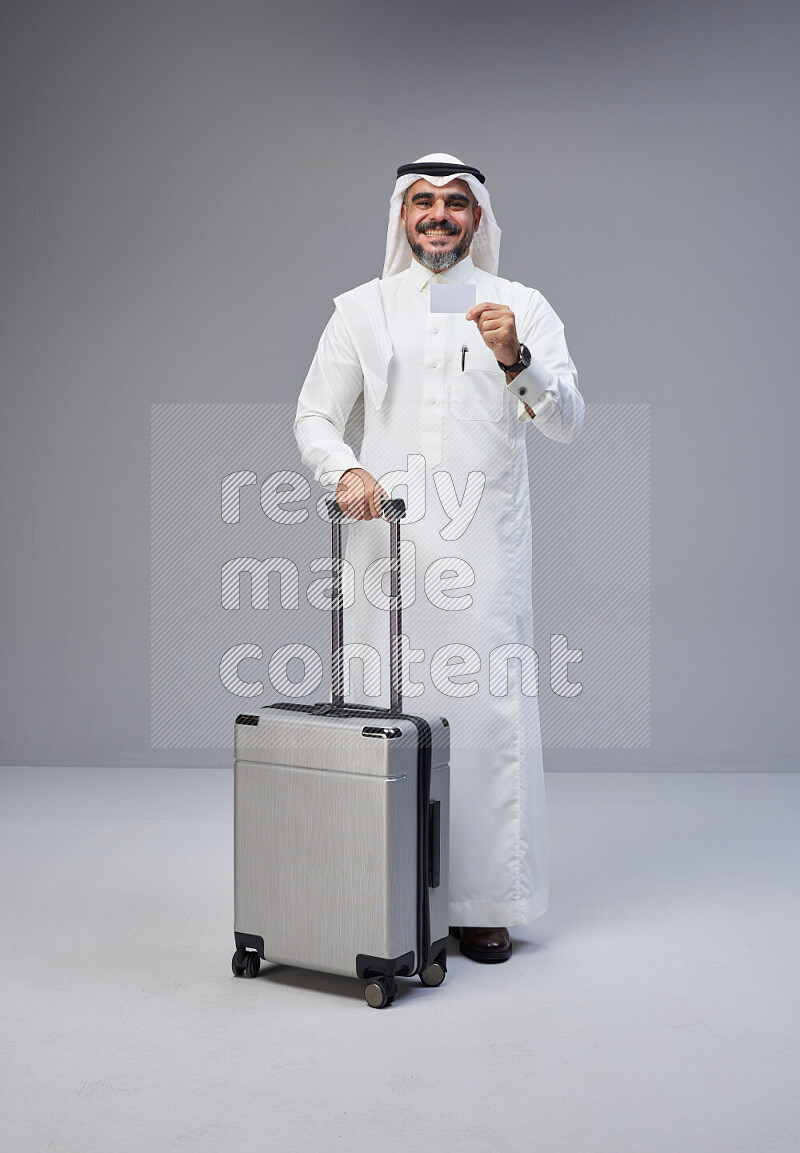 Saudi man wearing Thob and white Shomag standing holding Travel bag and ATM card on Gray background