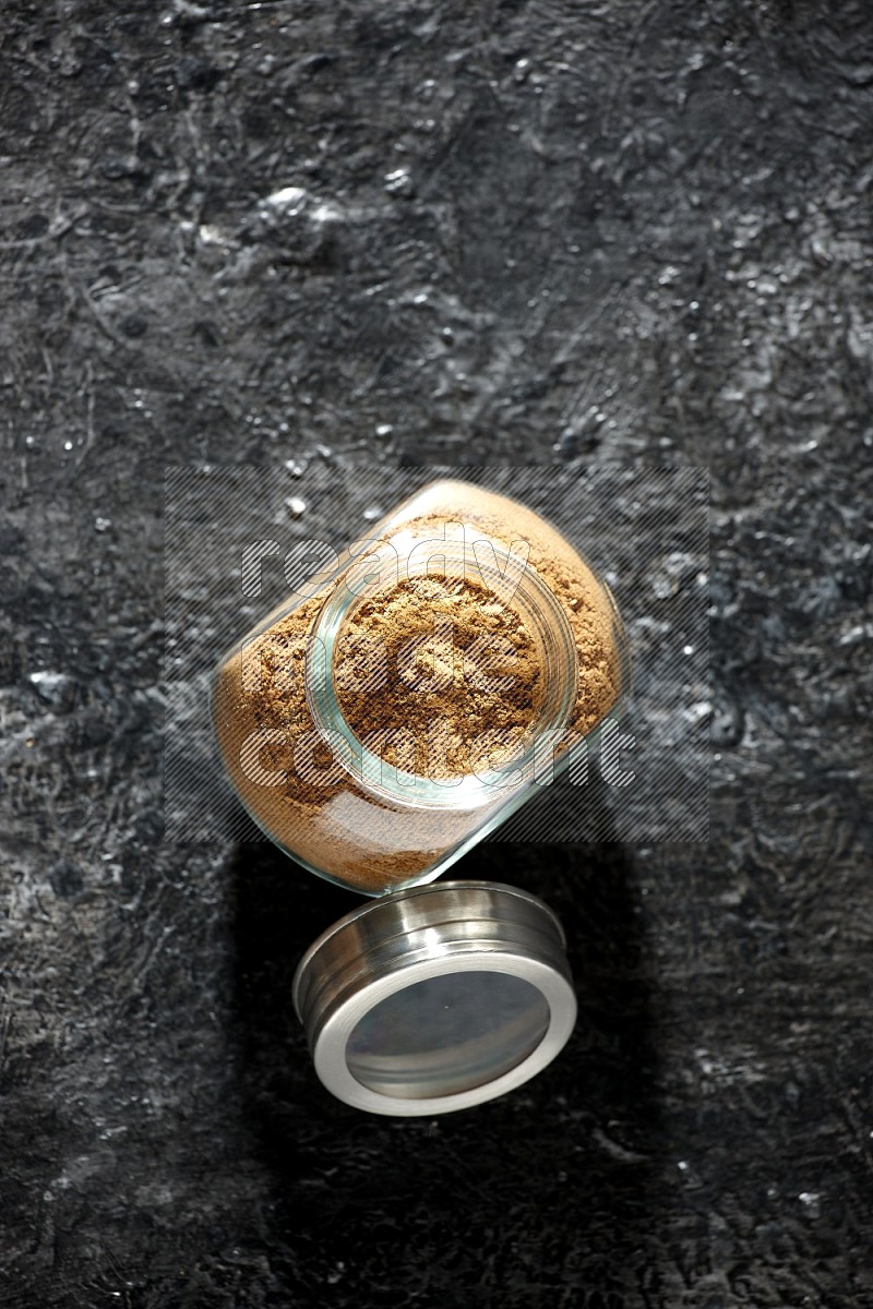 A glass spice jar full of allspice powder on a textured black flooring