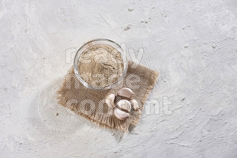 A glass bowl full of garlic powder placed on burlap fabric with garlic cloves on a textured white flooring
