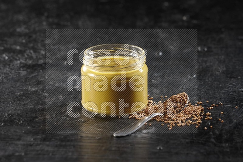 A glass jar full of mustard paste and mustard seeds in a metal spoon on a textured black flooring in different angles