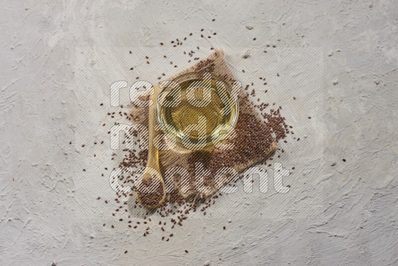 A glass bowl full of flaxseeds oil and wooden spoon full of flaxseeds with spread seeds on burlap fabric on a textured white flooring