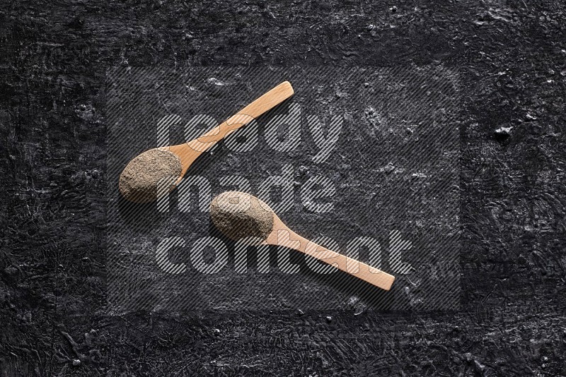 2 wooden spoons full of black pepper powder on a textured black flooring