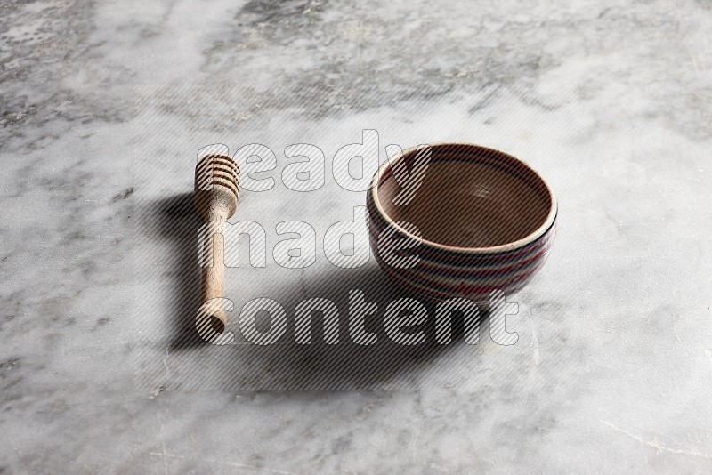 Multicolored Pottery bowl with wooden honey handle on the side with grey marble flooring, 45 degree angle