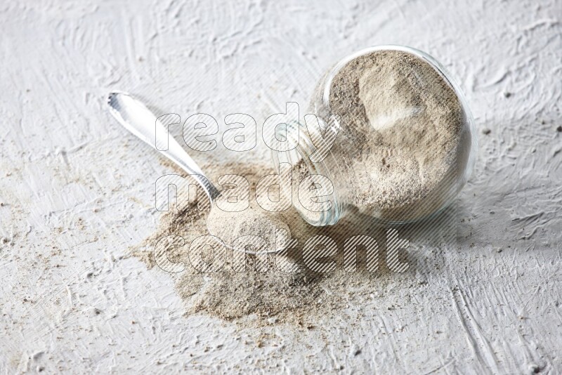 A flipped herbal glass jar and metal spoon full of white pepper powder with spilled powder on textured white flooring