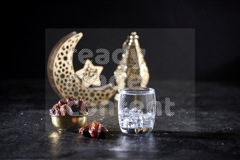 Dates in a metal bowl with water beside golden lanterns in a dark setup