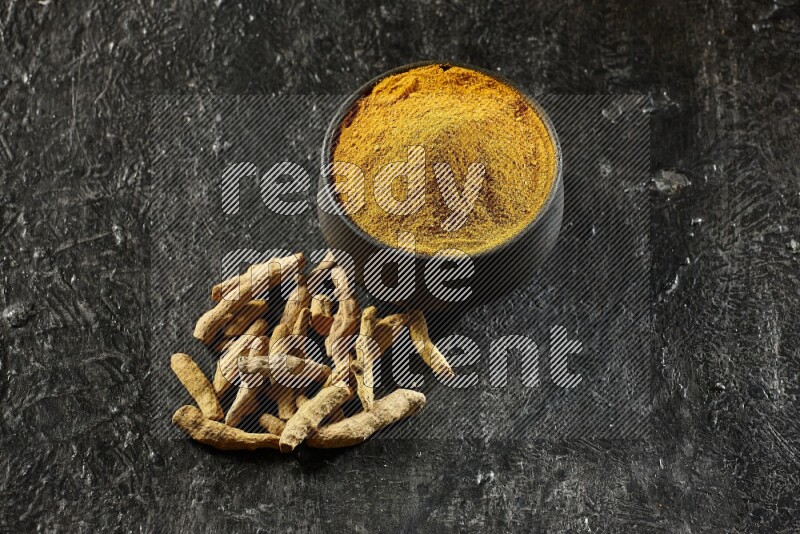 A black pottery bowl full of turmeric powder and dried turmeric whole fingers next of it on textured black flooring