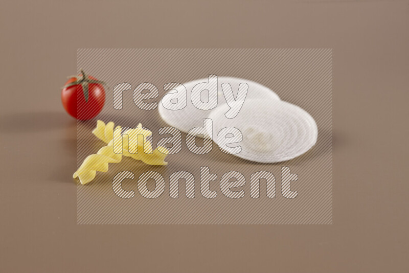 Raw pasta with different ingredients such as cherry tomatoes, garlic, onions, red chilis, black pepper, white pepper, bay laurel leaves, rosemary and cardamom on beige background
