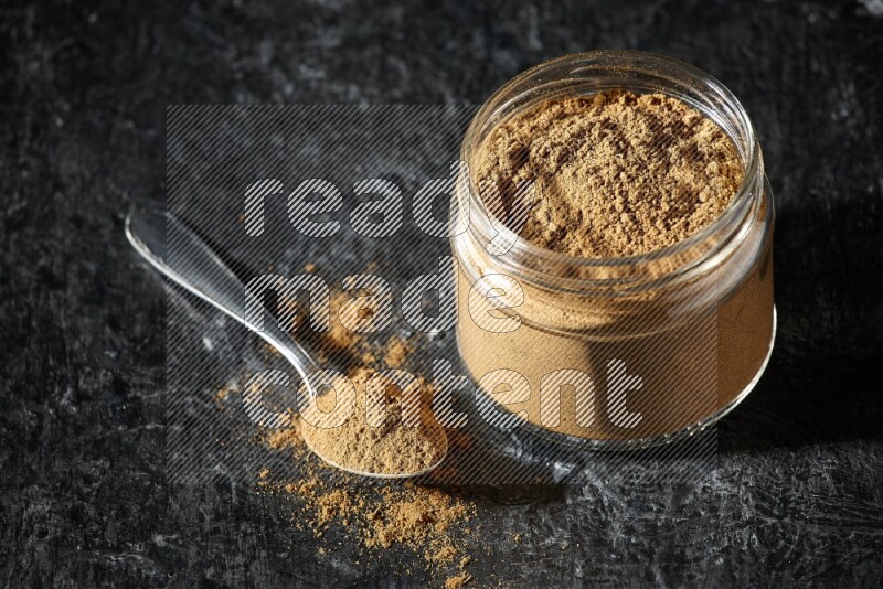 A glass jar and a metal spoon full of allspice powder on a textured black flooring