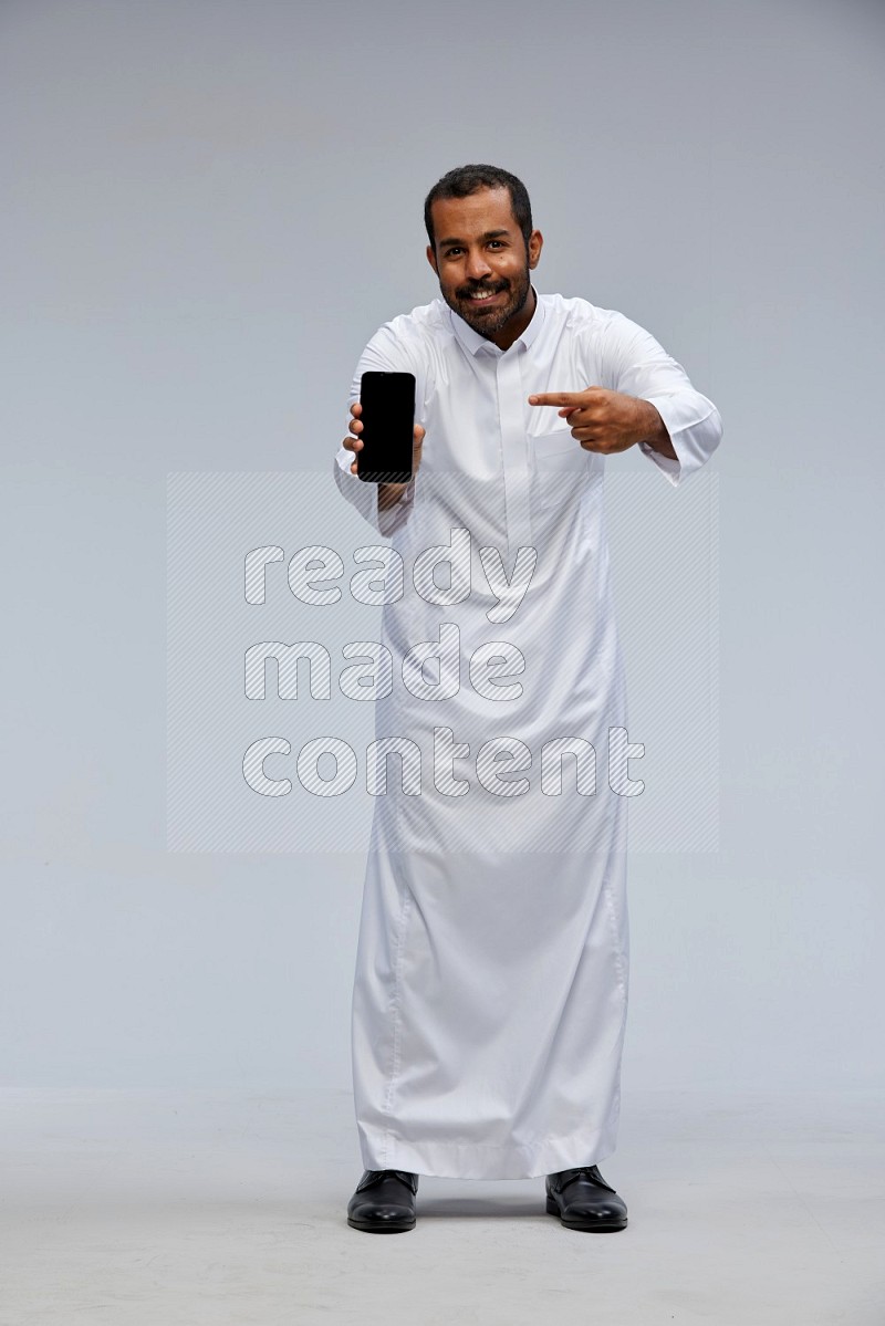 Saudi man wearing Thob standing showing phone to camera on Gray background