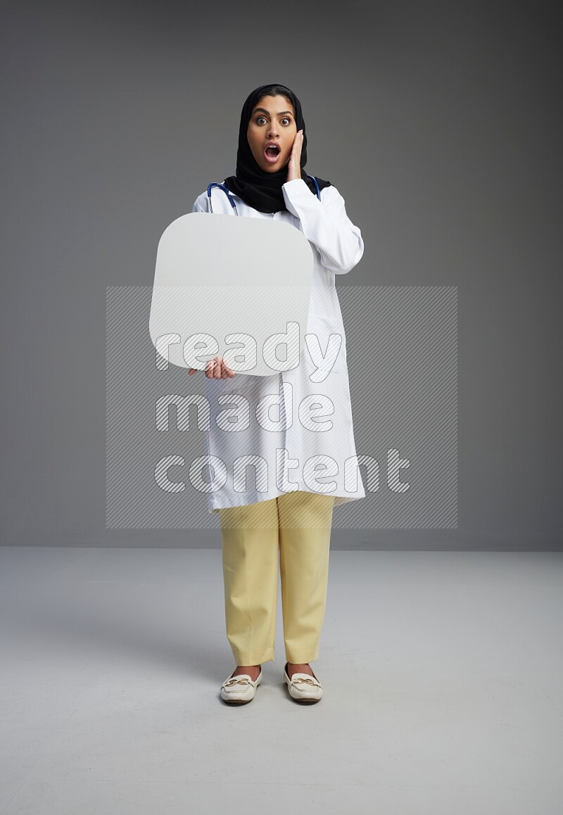 Saudi woman wearing lab coat with stethoscope standing holding social media sign on Gray background