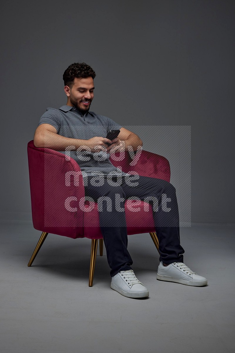 A man wearing casual and using his phone while sitting on a burgundy chair eye level on a gray background