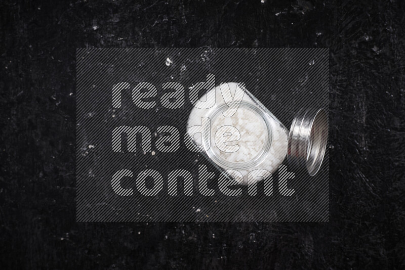 A glass jar full of coarse sea salt crystals on black background