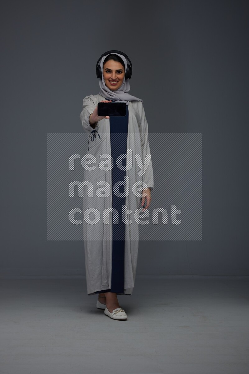 A Saudi woman wearing a light gray Abaya and head scarf setting on a dark grey chair and watching on the tablet eye level on a grey background