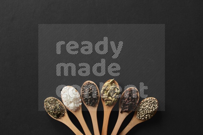 الفلفل الأبيض والقرنفل والهيل والملح والفلفل الأسود وريحان مجفف في معالق خشبية علي خلفية سوداء