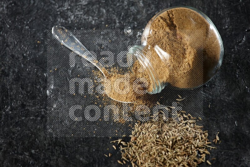 A flipped glass spice jar and a metal spoon full of cumin powder and powder spilled out with cumin seeds on a textured black flooring