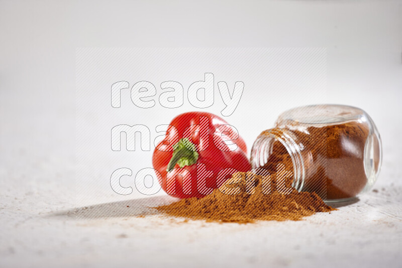 A glass jar full of ground paprika powder flipped with some spilling powder on white background