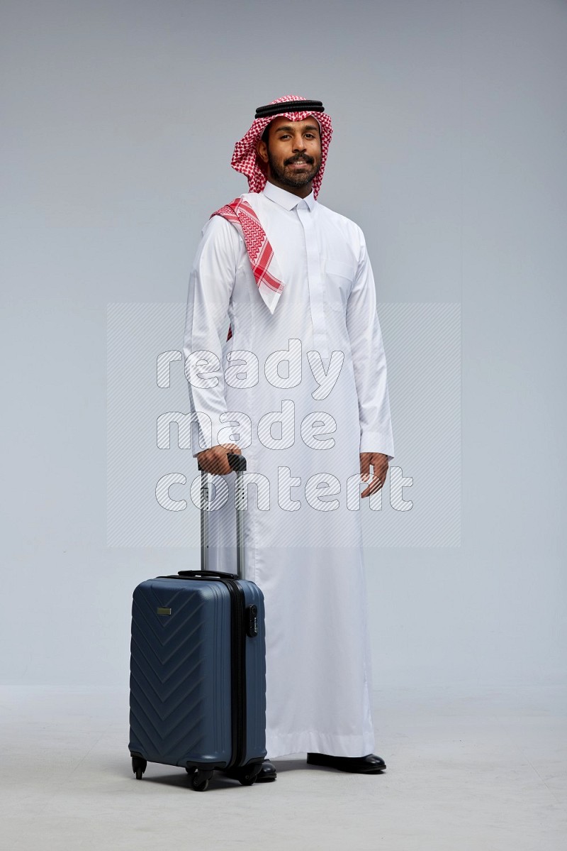 Saudi man wearing Thob and shomag standing holding Travel bag on Gray background