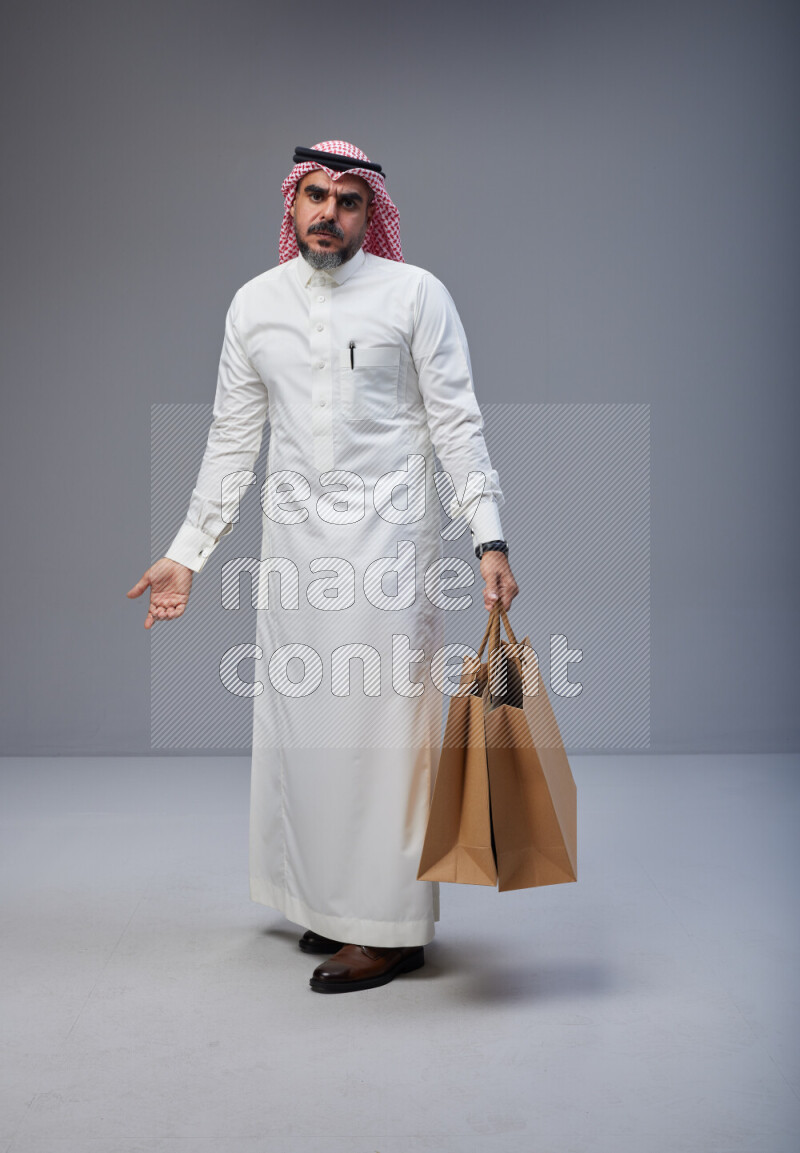 Saudi man Wearing Thob and red Shomag standing holding shopping bag on Gray background