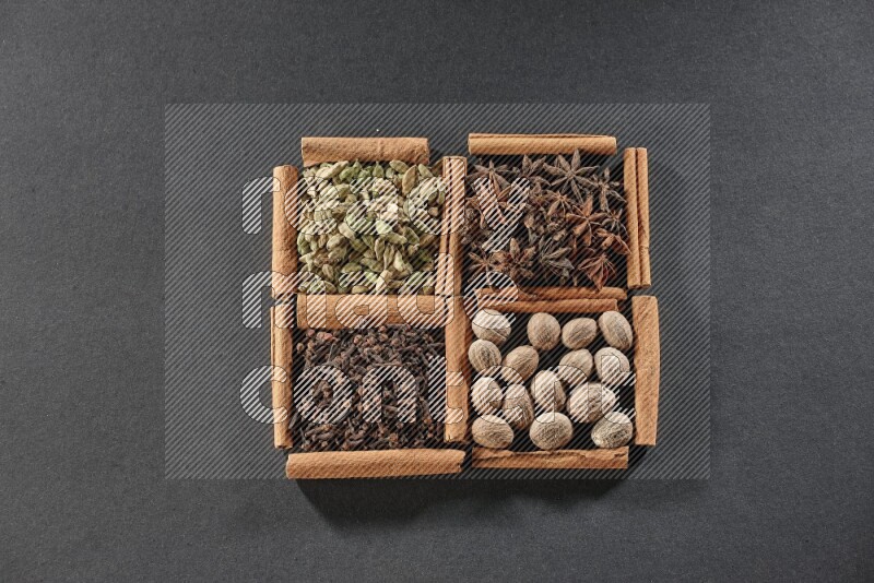 4 مربعات من عيدان القرفة ممتلئة بحب الهيل واليانسون النجمي وجوزة الطيب والقرنفل على أرضية سوداء