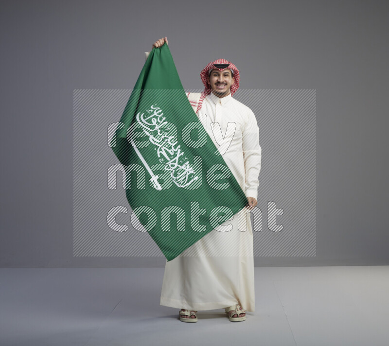 A saudi man standing wearing thob and red shomag holding big saudi flag on gray background