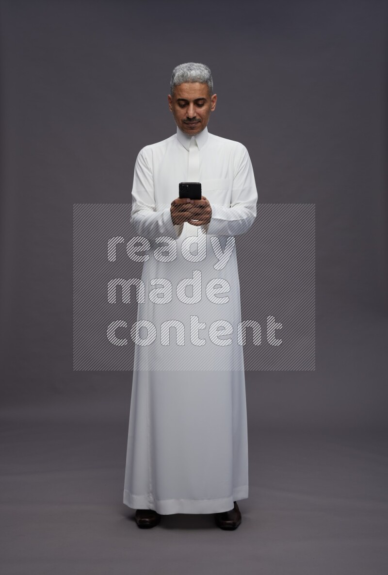 Saudi man wearing thob standing texting on phone on gray background