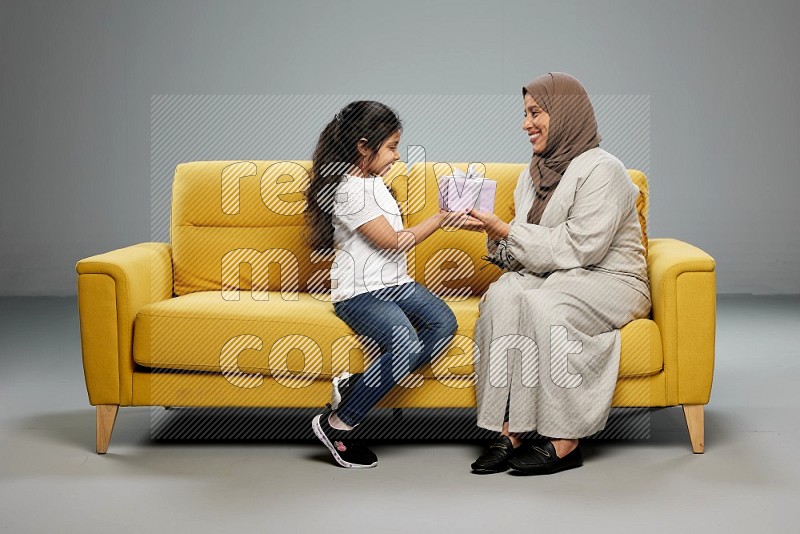 A girl sitting on a yellow sofa and giving a gift to her mother on gray background