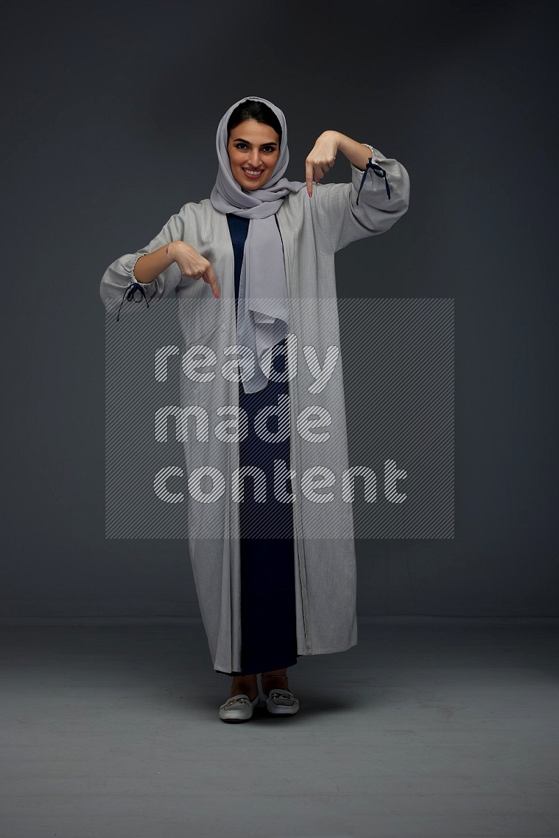 A Saudi woman wearing a light gray Abaya and head scarf standing and pointing in different directions eye level on a grey background