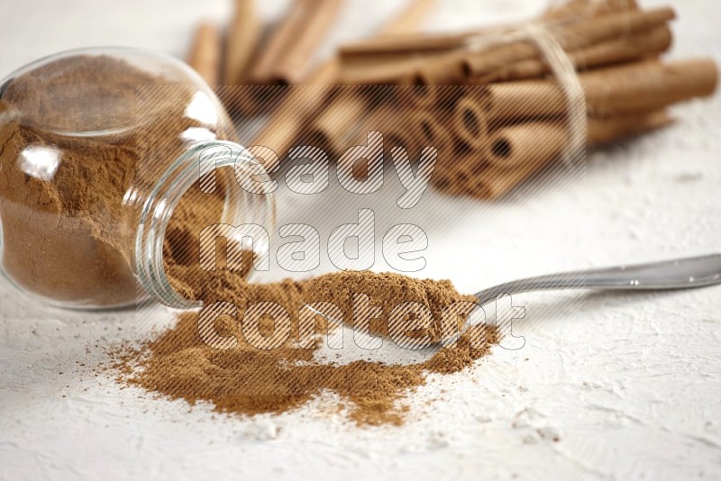 Flipped herbs glass jar full of cinnamon powder with a metal spoon full of powder and cinnamon sticks on a textured white background