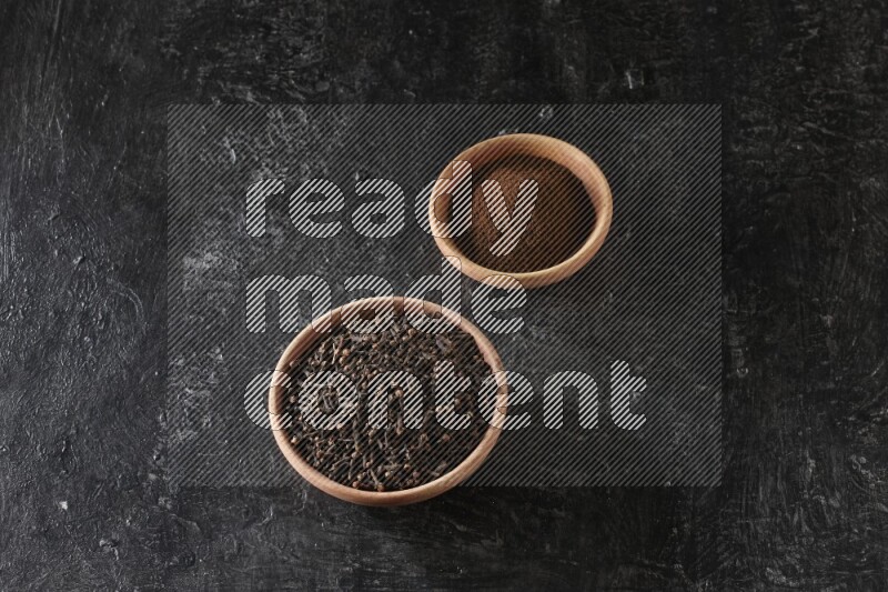 2 wooden bowls full of cloves powder and whole cloves on a textured black flooring