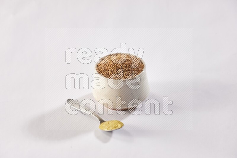 A beige pottery bowl full of mustard seeds and a metal spoon full of mustard paste on white flooring