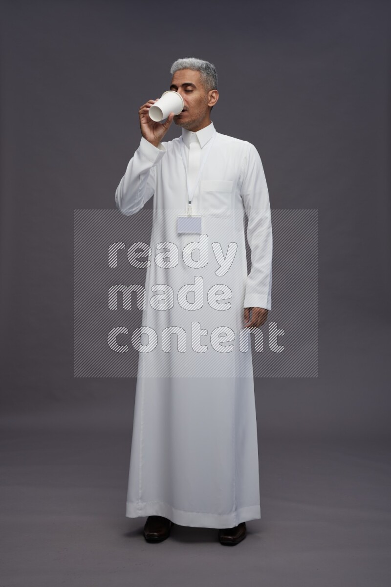 Saudi man wearing thob with neck strap employee badge standing holding paper cup on gray background