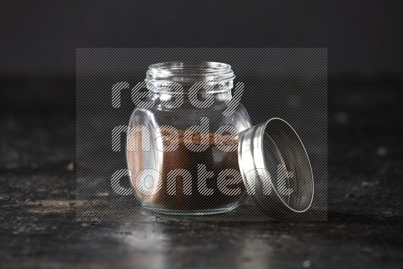 A glass spice jar full of cloves powder on textured black flooring