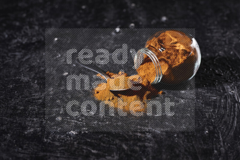 A glass jar full of ground paprika powder flipped with some spilling powder on black background