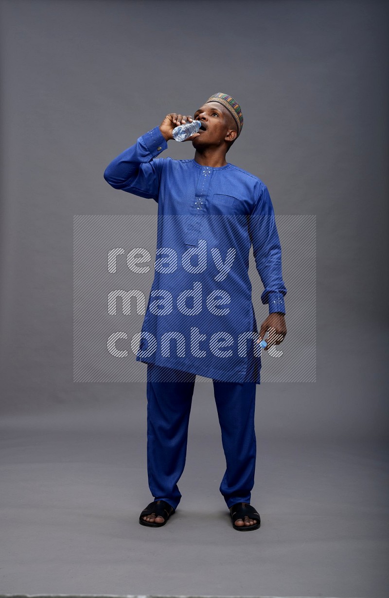 Man wearing Nigerian outfit standing drinking water on gray background