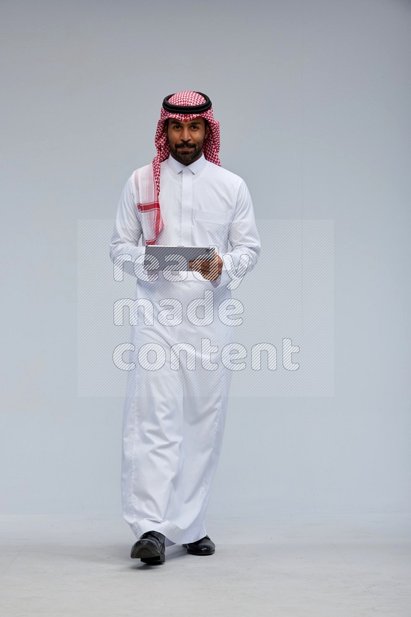 Saudi man Wearing Thob and shomag standing working on tablet on Gray background