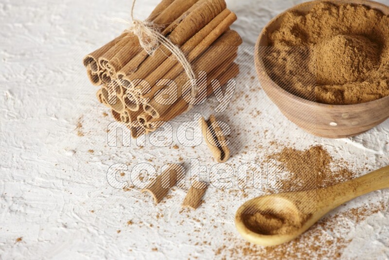 Cinnamon sticks stacked and bounded beside a wooden bowl full of cinnamon powder and a wooden spoon full of powder on white background