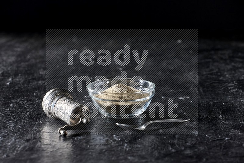 A glass bowl full of white pepper powder with pepper beads, a metal grinder and a metal spoon on textured black flooring