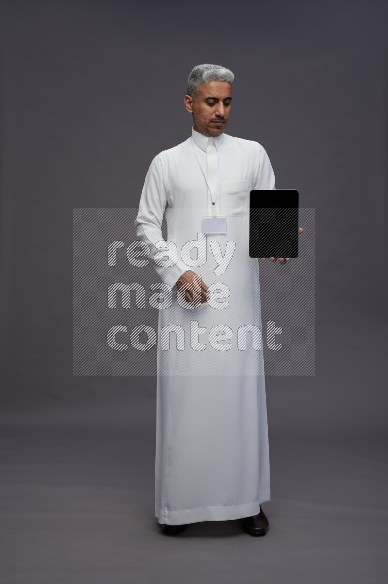 Saudi man wearing thob with neck strap employee badge standing showing tablet to camera on gray background