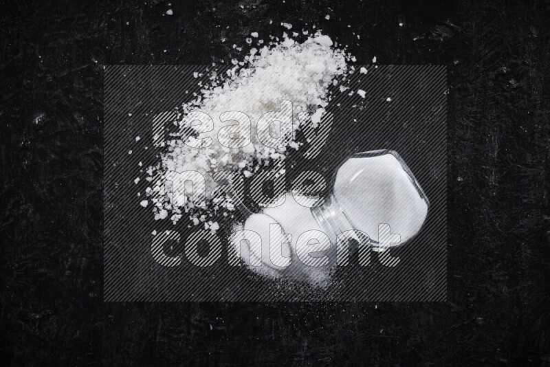 A glass jar full of table salt with some sea salt crystals beside it on a black background