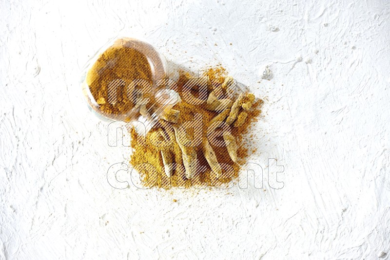 A flipped glass spice jar full of turmeric powder and powder spilled out of it with dried whole fingers on textured white flooring