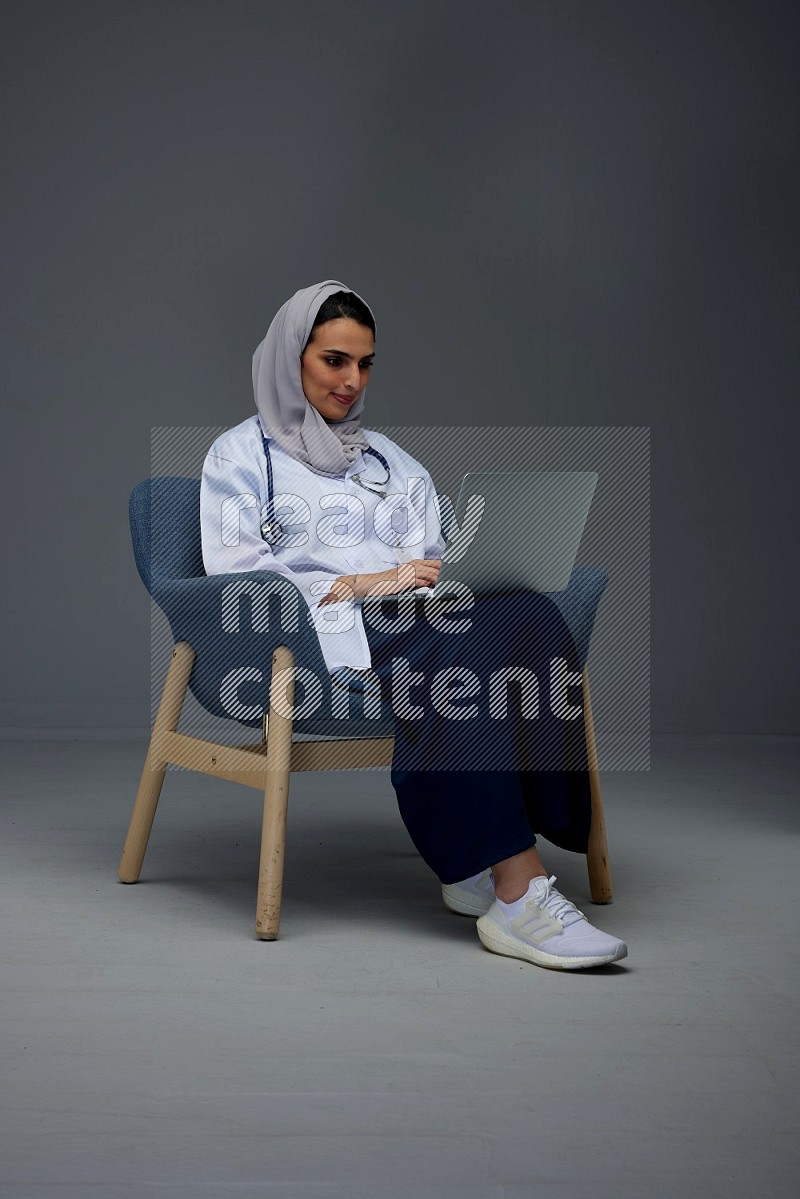 A doctor wearing a light gray head scarf sitting on a dark grey chair and using a laptop eye level on a grey background