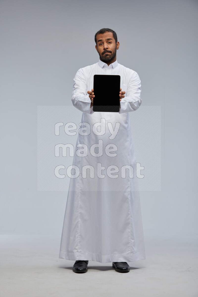 Saudi man wearing Thob standing showing tablet to camera on Gray background