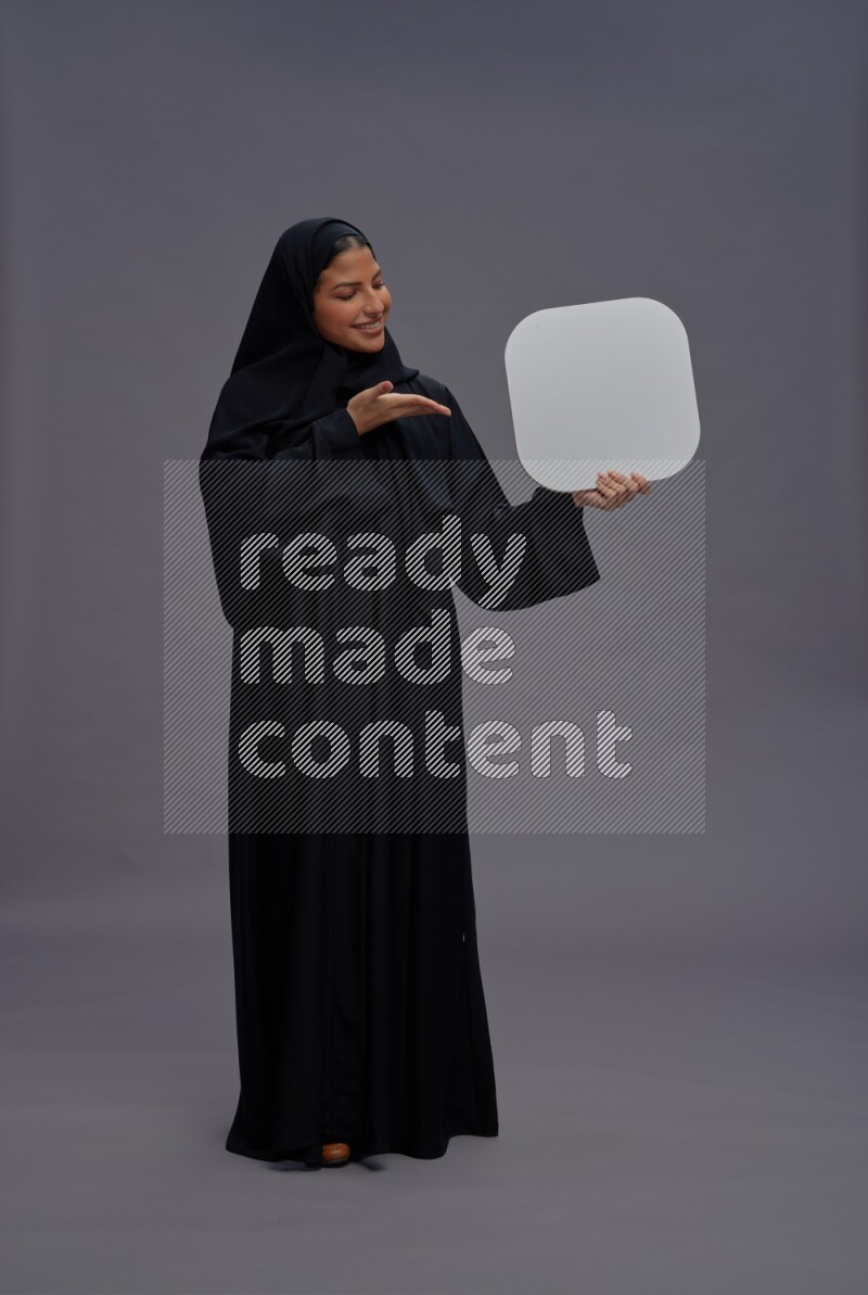 Saudi woman wearing Abaya standing holding social media sign on gray background