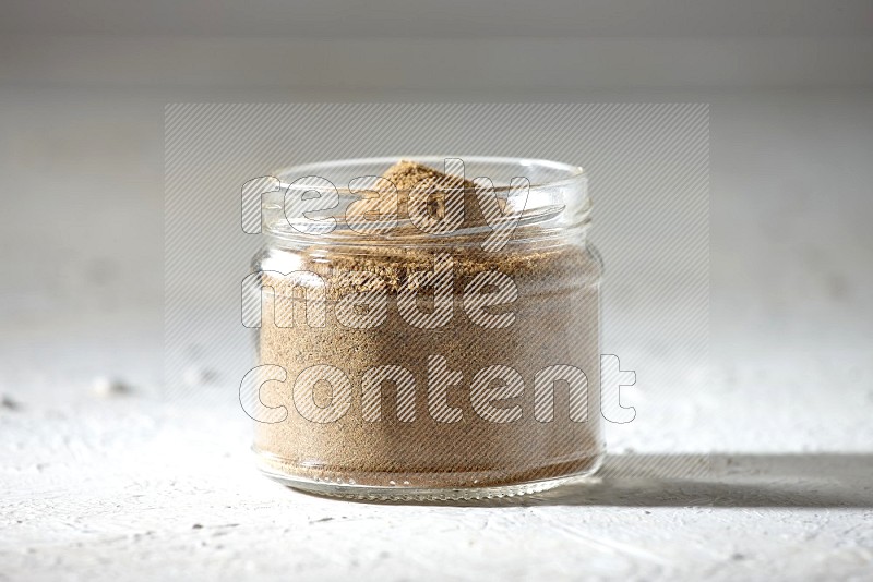 A glass jar full of cumin powder on textured white flooring